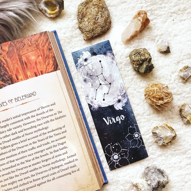 May include: A black and white bookmark with a watercolor design of the Virgo constellation and flowers. The bookmark has the word "Virgo" printed in white on a black background.