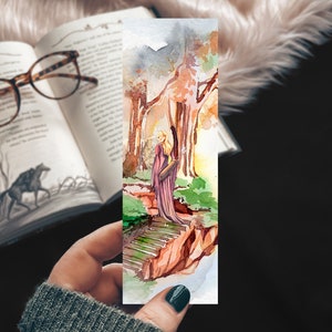 May include: A watercolour bookmark depicting a woman in a purple robe standing on a stone path in a forest. The trees are painted in shades of brown, green, and orange. The background is a soft, watercolour wash of blue and white.