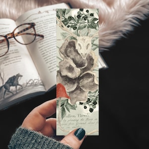 May include: A rectangular bookmark featuring a watercolor illustration of a deer skull, dark mushrooms, green foliage, and a single red mushroom. The bottom section includes printed text that reads Saffron Flower and additional botanical descriptions.