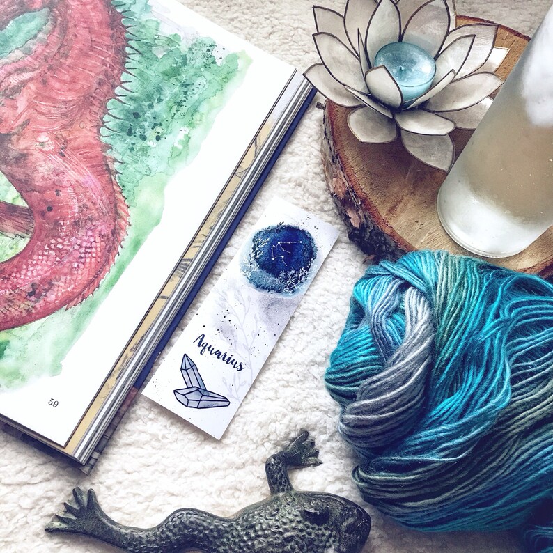 May include: A watercolor bookmark with a blue and white illustration of the Aquarius constellation and the word "Aquarius" written in black ink. The bookmark is lying on a white fuzzy surface next to a book, a frog figurine, a yarn ball, a wooden log, and a white lotus flower candle holder.