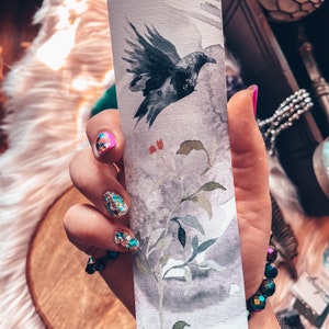May include: A hand holding a bookmark featuring a watercolour illustration of a black bird in flight above a branch with green leaves. The background is a light grey with a watercolour wash. The hand has glitter nail polish.
