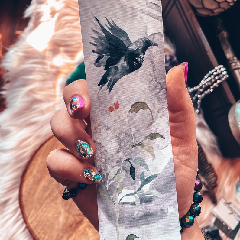 May include: A hand holding a bookmark featuring a watercolour illustration of a black bird in flight above a branch with green leaves. The background is a light grey with a watercolour wash. The hand has glitter nail polish.