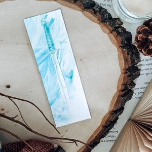 May include: A blue and white watercolor bookmark featuring a detailed illustration of a sword.