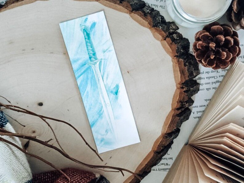 May include: A blue and white watercolor bookmark featuring a detailed illustration of a sword.