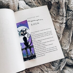 Peut inclure: Un chat noir aux yeux verts est assis sur un poteau de cl&ocirc;ture devant une lune violette et bleue. Le chat regarde vers la droite. L'image est tir&eacute;e d'un livre avec le texte "CHAPTER ONE RHEN" sur la page.