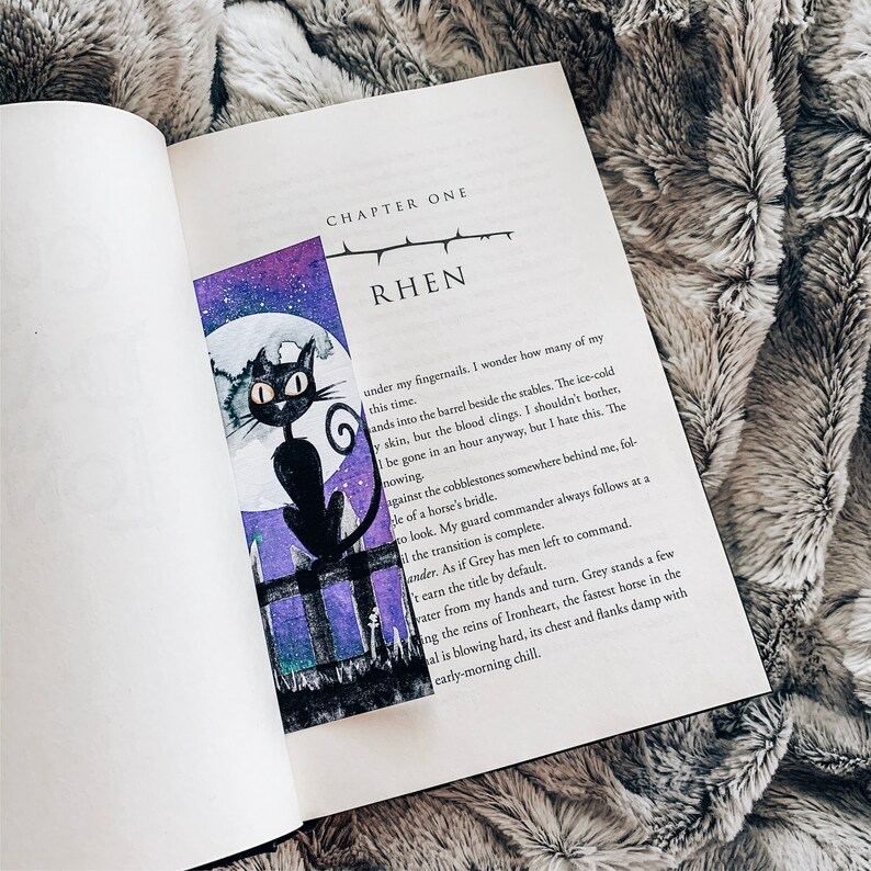 Peut inclure: Un chat noir aux yeux verts est assis sur un poteau de cl&ocirc;ture devant une lune violette et bleue. Le chat regarde vers la droite. L'image est tir&eacute;e d'un livre avec le texte "CHAPTER ONE RHEN" sur la page.