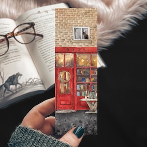 May include: A watercolor illustration of a red brick building with a red door and a window with a cat looking out. The building has a sign that says "BOOK." There are lights strung above the door and bookshelves inside the building.