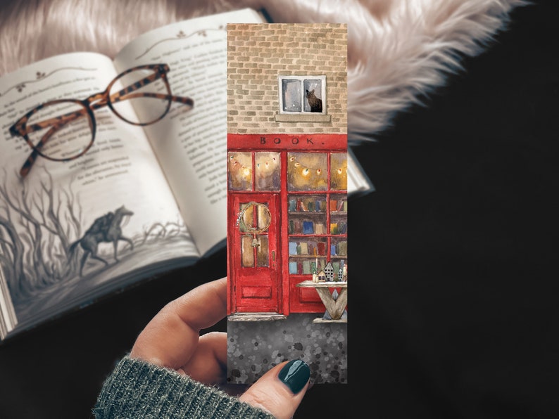 May include: A watercolor illustration of a red brick building with a red door and a window with a cat looking out. The building has a sign that says "BOOK." There are lights strung above the door and bookshelves inside the building.