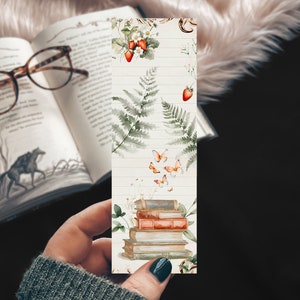 May include: A decorative bookmark featuring watercolor illustrations of strawberries, ferns, butterflies, and a stack of books. The bookmark has a light wood grain background and is held in a person's hand, with an open book and glasses in the background.