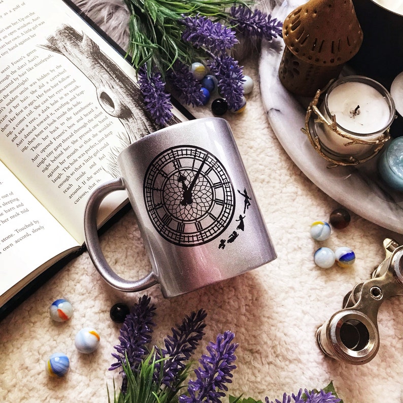 May include: A silver mug with a black and white clock face and silhouettes of Peter Pan and the Lost Boys flying around it. The mug is sitting on a white surface with purple flowers and marbles.