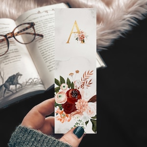 May include: A gold letter A bookmark with a floral design. The flowers are red, pink, and white. The bookmark is on a marble background.