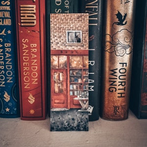 May include: A close-up of a book-themed bookmark with a watercolor illustration of a brick building with a shop front. The shop has a sign that reads "BOOK" and a window with a black cat inside. The bookmark is surrounded by a row of books with colorful spines.