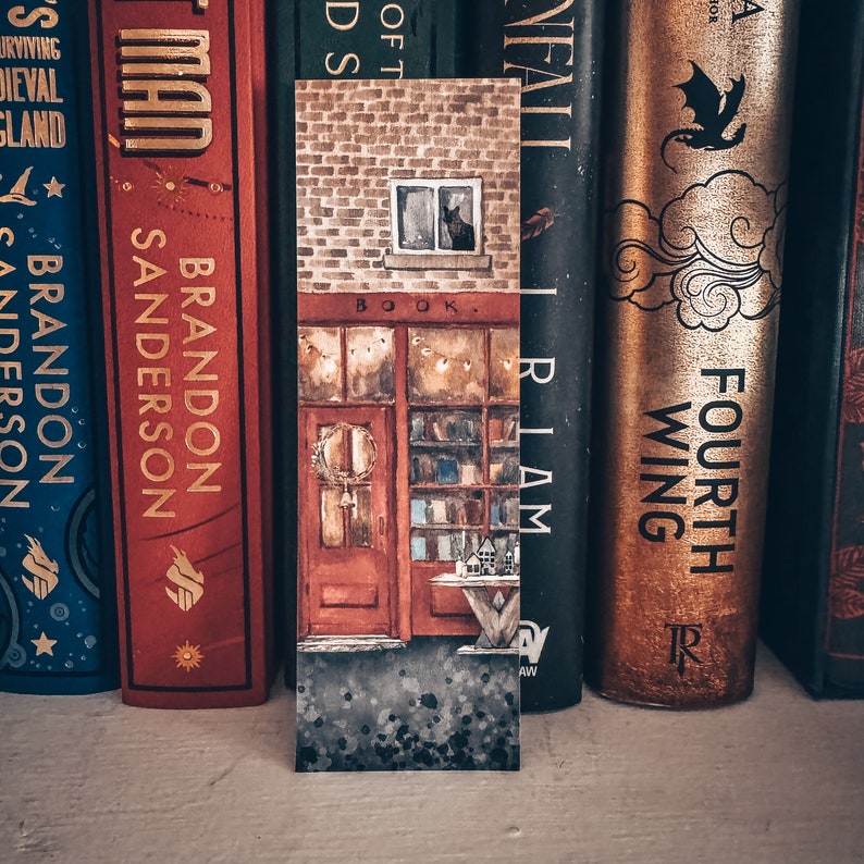 May include: A close-up of a book-themed bookmark with a watercolor illustration of a brick building with a shop front. The shop has a sign that reads "BOOK" and a window with a black cat inside. The bookmark is surrounded by a row of books with colorful spines.