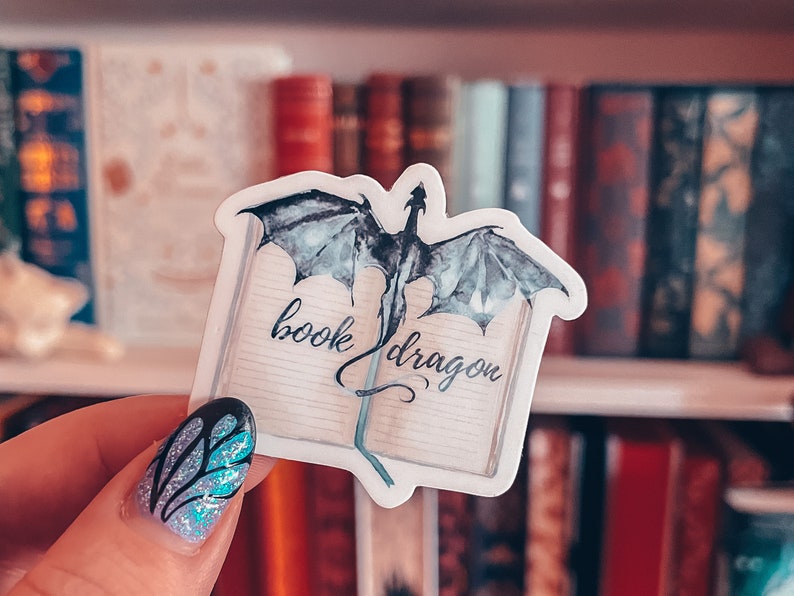 May include: A white sticker featuring a watercolour illustration of a dragon emerging from an open book. The book has the words "book dragon" written on it.