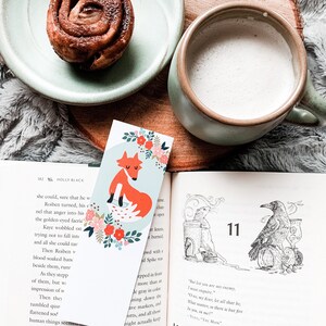 May include: A close-up of a book with a bookmark featuring a fox illustration. The bookmark is on a light blue background with red, white, and green floral accents. The book is open to a page with a black and white illustration of a crow.