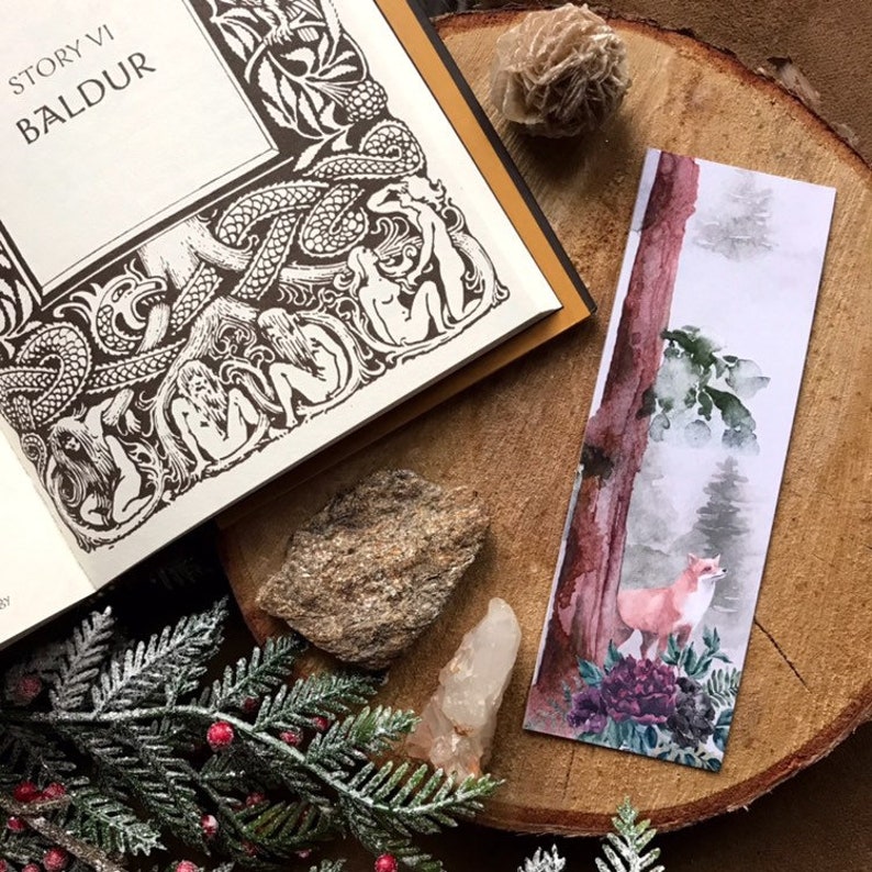 May include: A watercolor bookmark featuring a red fox standing in a forest scene. The fox is in the foreground, with a tall tree trunk and lush green foliage behind it. The background is a soft, misty blue.