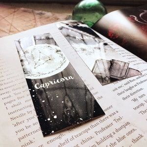 May include: A black and white Capricorn zodiac sign bookmark with a constellation design. The bookmark features the word "Capricorn" in white script. The bookmark is on top of an open book.