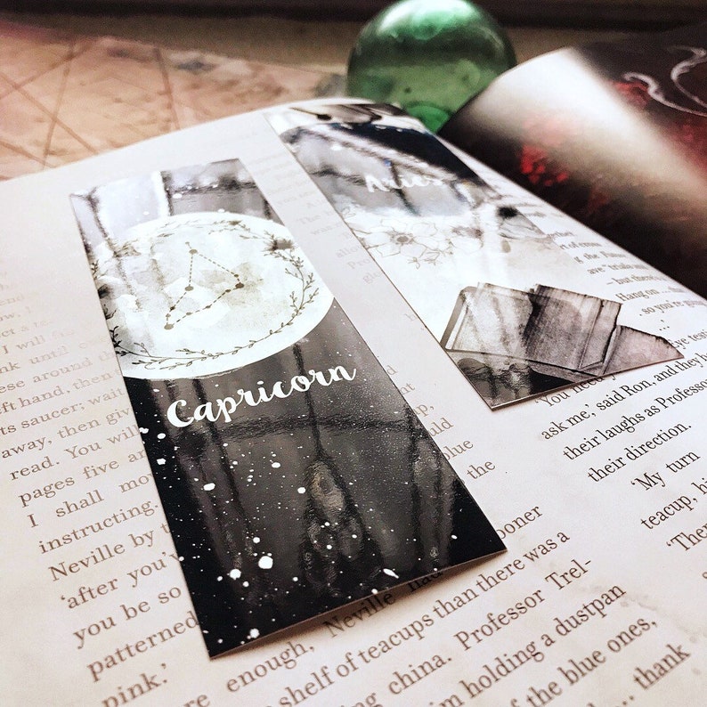 May include: A black and white Capricorn zodiac sign bookmark with a constellation design. The bookmark features the word "Capricorn" in white script. The bookmark is on top of an open book.