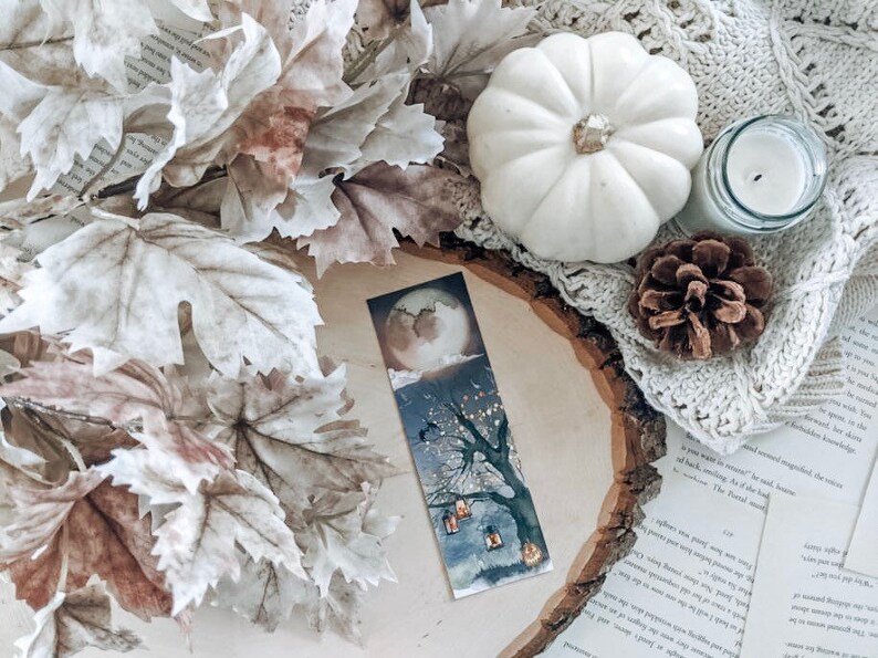 Peut inclure: Une citrouille blanche, une pomme de pin brune, une bougie blanche et un livre avec un marque-page repr&eacute;sentant une illustration aquarelle d'un arbre avec des lanternes sous une lune croissante. Le marque-page est sur une tranche de bois.