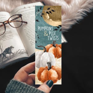 May include: A watercolor bookmark featuring a teal background with the text "Pumpkins & Plot Twists." The bookmark showcases a white pumpkin and two orange pumpkins. A gold moon and black leaves are also depicted.