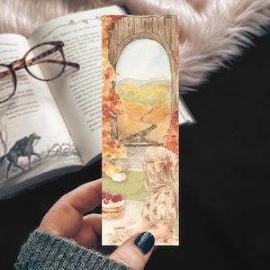 May include: A rectangular bookmark featuring a watercolor illustration of a person with long hair sitting at a table with a cake, looking through a stone archway at a landscape with autumn trees, rolling hills, and a winding path in warm tones.
