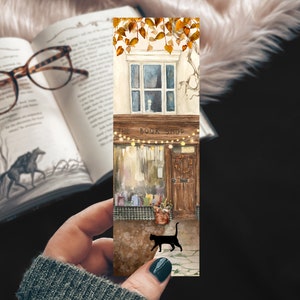 May include: A watercolor bookmark featuring a quaint bookstore with a black cat walking in front. The shop has a sign that reads "The Book Shop" with string lights above the door. The building is decorated with fall leaves.
