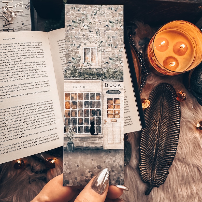 Peut inclure: Peinture &agrave; l'aquarelle d'un petit b&acirc;timent en briques avec une enseigne qui indique "Cafe" et "Book". Le b&acirc;timent a une grande fen&ecirc;tre avec un chat noir assis dans la fen&ecirc;tre. Le b&acirc;timent est peint dans des tons de brun, gris et vert.