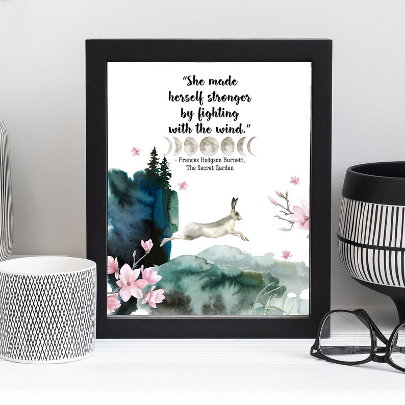 Puede incluir: Una impresi&oacute;n enmarcada en negro con un fondo blanco que presenta una cita de Frances Hodgson Burnett's The Secret Garden. La cita dice "She made herself stronger by fighting with the wind." La impresi&oacute;n tambi&eacute;n presenta una ilustraci&oacute;n de acuarela de una liebre saltando sobre una colina con un dise&ntilde;o de fase lunar arriba.