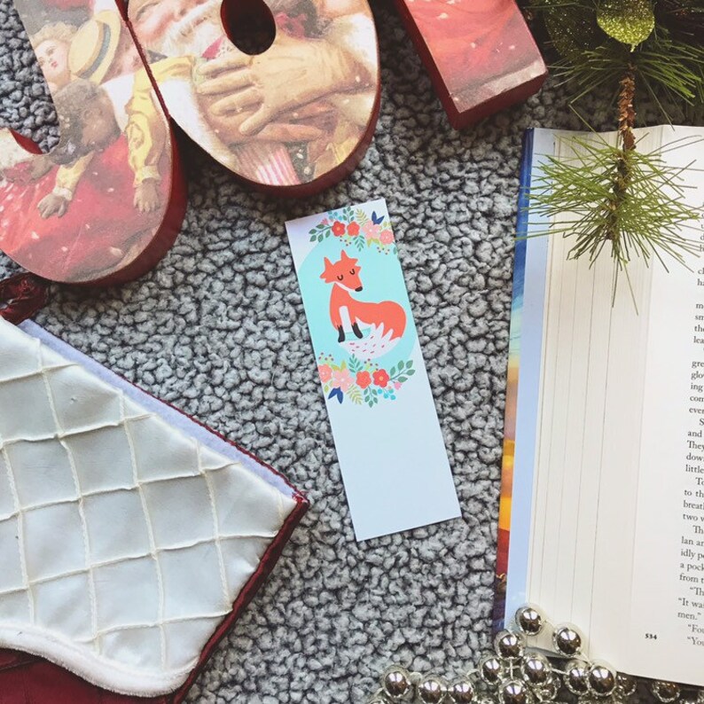 May include: A bookmark featuring a cute fox illustration with a floral design. The fox is sitting on a blue background with a white border. The bookmark is perfect for book lovers and animal enthusiasts.
