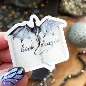 May include: A white sticker featuring a watercolour illustration of a black dragon emerging from an open book. The text "book dragon" is written in cursive on the book.