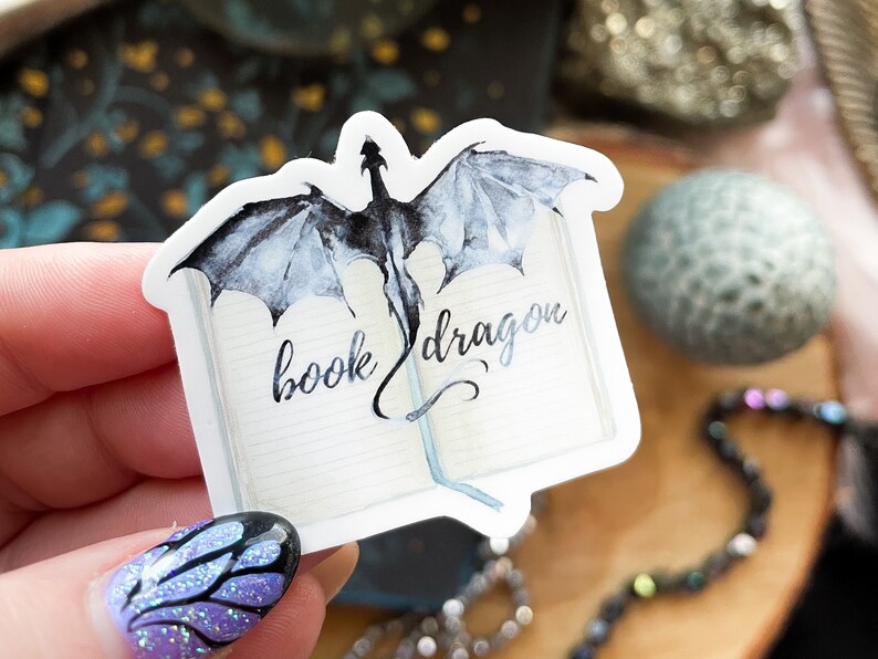 May include: A white sticker featuring a watercolour illustration of a black dragon emerging from an open book. The text "book dragon" is written in cursive on the book.