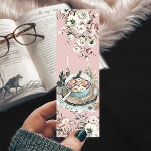 May include: A pink bookmark with a watercolor illustration of a bowl of mulled cider with cinnamon sticks, oranges, and apples. The bowl is on a wooden cutting board with a spoon. There are white flowers and greenery around the bowl.