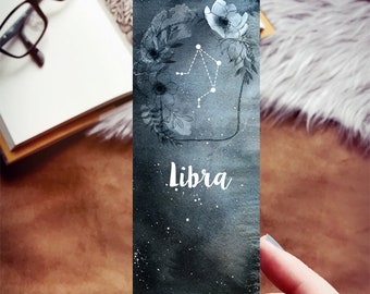 May include: A watercolor bookmark featuring a Libra zodiac design. The bookmark has a dark blue and gray background with white floral accents and the Libra constellation. The word "Libra" is written in white script.
