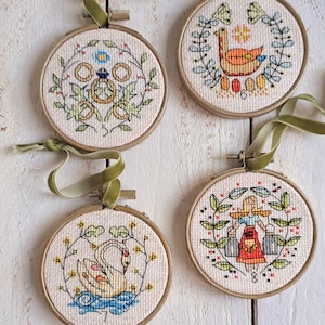 May include: Four round embroidery hoops featuring different designs. The hoops are made of wood and have a white background. The designs include a bird, a swan, a woman carrying buckets, and a ring. The hoops are all hanging from a white string.