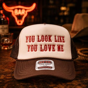 May include: A brown and white trucker hat featuring the embroidered red text "YOU LOOK LIKE YOU LOVE ME" on the front foam panel. The hat has a brown mesh back, a brown brim, and a sticker on the brim that reads "OTTO WORLD FAMOUS FOAM FRONT OSFM".