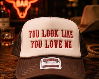 May include: A brown and white trucker hat featuring the embroidered red text "YOU LOOK LIKE YOU LOVE ME" on the front foam panel. The hat has a brown mesh back, a brown brim, and a sticker on the brim that reads "OTTO WORLD FAMOUS FOAM FRONT OSFM".
