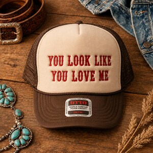 May include: A brown and cream trucker hat featuring the red embroidered text YOU LOOK LIKE YOU LOVE ME on the front foam panel. The hat includes a brown mesh back, a brown brim, and a white sticker on the brim with the text OTTO WORLD FAMOUS FOAM FRONT.
