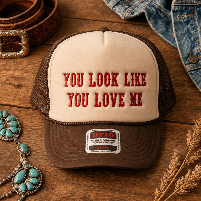 May include: A brown and cream trucker hat featuring the red embroidered text YOU LOOK LIKE YOU LOVE ME on the front foam panel. The hat includes a brown mesh back, a brown brim, and a white sticker on the brim with the text OTTO WORLD FAMOUS FOAM FRONT.