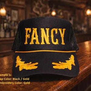 May include: A black trucker cap featuring the word FANCY embroidered in gold thread across the front panel. The cap includes a gold cord trim above the brim and gold leaf embroidery on the brim. The sides and back are made of black mesh material.