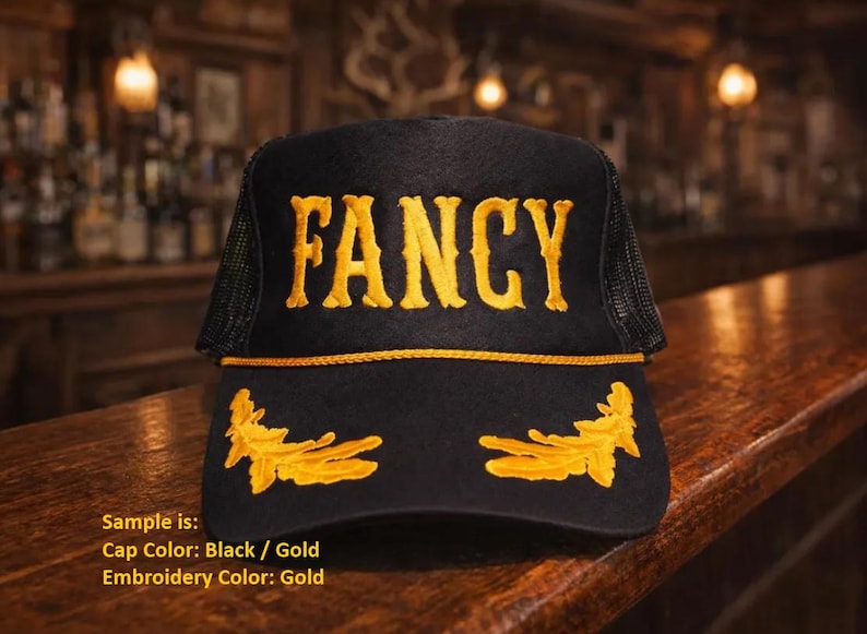 May include: A black trucker cap featuring the word FANCY embroidered in gold thread across the front panel. The cap includes a gold cord trim above the brim and gold leaf embroidery on the brim. The sides and back are made of black mesh material.
