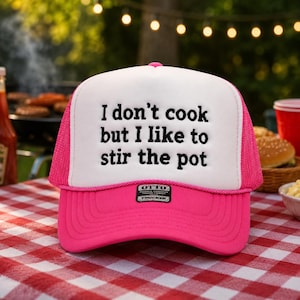 May include: A pink and white trucker hat with the text "I don't cook but I like to stir the pot." The hat has a pink brim and mesh back. The hat is sitting on a red and white checkered tablecloth.
