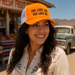 May include: A bright orange trucker hat with a white mesh back and a curved brim. The front panel features the black text "YOU LOOK LIKE YOU LOVE ME" in a bold, sans-serif font. The hat has a structured crown and an adjustable snapback closure.