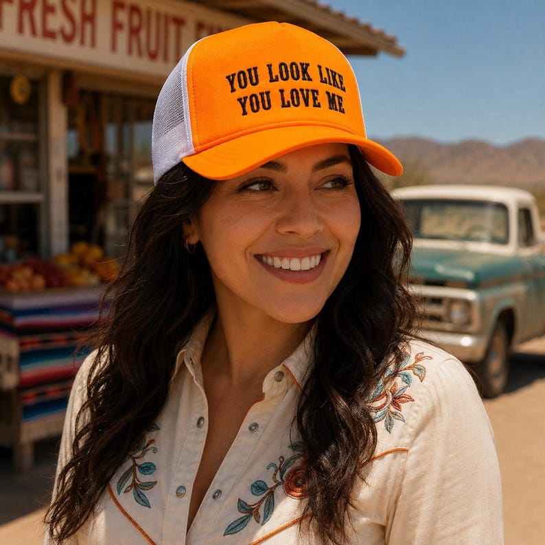 May include: A bright orange trucker hat with a white mesh back and a curved brim. The front panel features the black text "YOU LOOK LIKE YOU LOVE ME" in a bold, sans-serif font. The hat has a structured crown and an adjustable snapback closure.