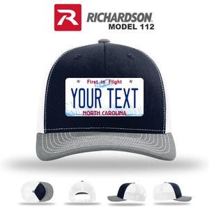 May include: A navy blue and white trucker hat with a gray brim. The hat features a rectangular patch with the text "YOUR TEXT" and "NORTH CAROLINA" on it. The patch also has the text "First in Flight".