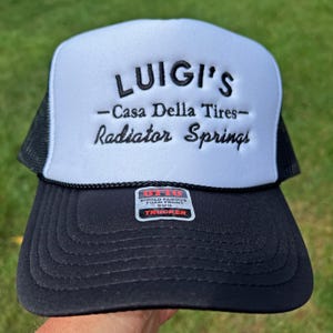 May include: A white and black trucker hat with the text "LUIGI'S -Casa Della Tires- Radiator Springs" embroidered in black on the front. The hat has a black mesh back and a black bill with a red and white "Otto World Famous Foam Front 0.5FM Trucker" label.