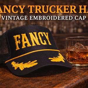 May include: A black trucker hat with a mesh back, featuring the word FANCY embroidered in yellow on the front panel, yellow decorative embroidery on the brim, and a yellow cord trim. Next to it sits a glass of amber liquid with ice cubes.