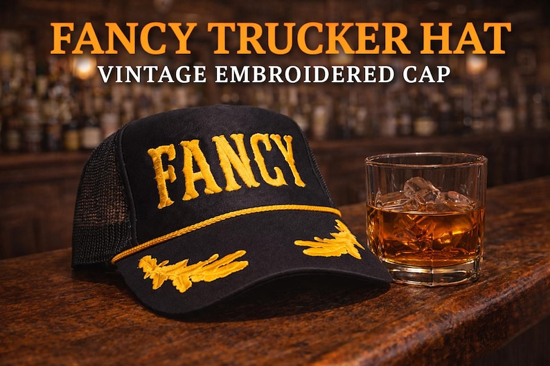 May include: A black trucker hat with a mesh back, featuring the word FANCY embroidered in yellow on the front panel, yellow decorative embroidery on the brim, and a yellow cord trim. Next to it sits a glass of amber liquid with ice cubes.