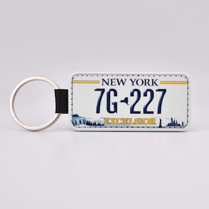 May include: White keychain with a New York license plate design. The plate reads "NEW YORK 7G-227 EXCELSIOR".