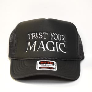 May include: A black trucker hat with a mesh back. The front of the hat has white embroidery that reads "TRUST YOUR MAGIC".