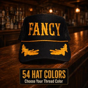 May include: A black trucker hat featuring the word FANCY embroidered in gold thread across the front panel, with gold laurel leaf embroidery on the brim and a gold cord trim. A rectangular graphic below displays the text 54 HAT COLORS CHOOSE YOUR THREAD COLOR.